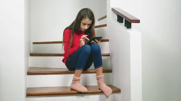 Vacker flicka i en röd tröja och terry strumpor och jeans, sitter på trappan, använder tabletten — Stockfoto