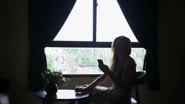 Silhouette. Mädchen sitzt auf einem Stuhl vor dem Fenster, benutzt ihr Handy und trinkt Wasser aus einem Glas — Stockfoto