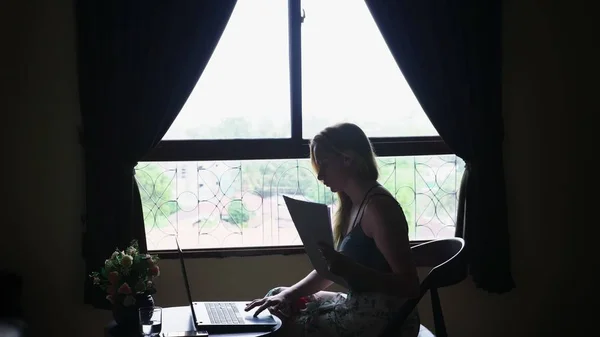 Σιλουέτα. κορίτσι που κάθεται σε μια καρέκλα από το παράθυρο, χρησιμοποιεί το φορητό υπολογιστή, κοιτάζοντας μέσα από έγγραφα σε χαρτί — Φωτογραφία Αρχείου