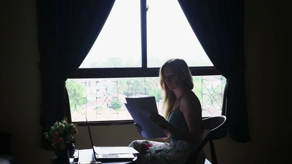 Silueta. chica se sienta en una silla junto a la ventana, utiliza su computadora portátil, mirando a través de documentos de papel — Foto de Stock
