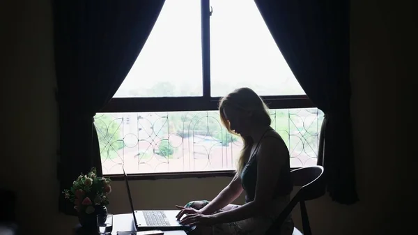 Σιλουέτα. κορίτσι να κάθεται σε μια καρέκλα από το παράθυρο, να χρησιμοποιεί το φορητό υπολογιστή. — Φωτογραφία Αρχείου