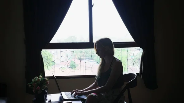 Silhouette. Mädchen sitzt auf einem Stuhl am Fenster, benutzt ihren Laptop. — Stockfoto