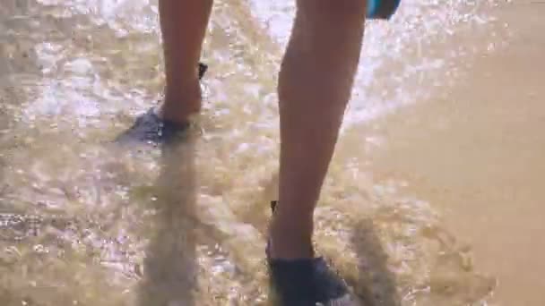 Primer plano de las piernas en zapatos de baño negro caminando a lo largo de la costa de la playa en agua de mar. hombre sostiene tabla de natación — Vídeo de stock