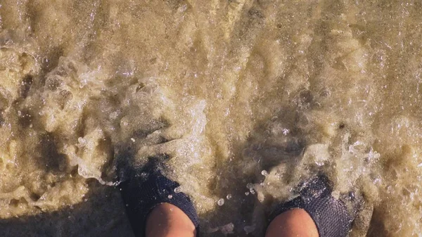 Close-up das pernas em sapatos de natação pretos andando ao longo da costa da praia em água do mar. homem detém natação bordo — Fotografia de Stock