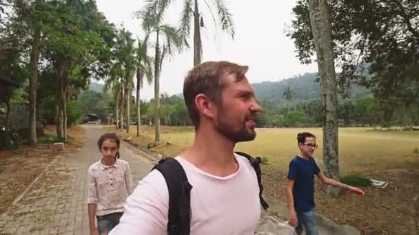 Eine glückliche Familie von Bloggern mit Kameras nimmt einen Blog mit einer Action-Kamera auf, die durch einen tropischen Park spaziert. — Stockvideo