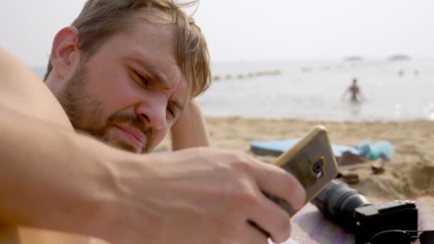 Jovem deita-se na praia à beira-mar e usa seu telefone — Vídeo de Stock