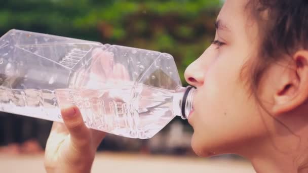 Gros plan. une fille boit de l'eau propre d'une bouteille en plastique sur un fond de feuilles vertes — Video