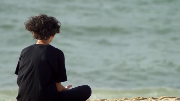 Ragazzo, adolescente dai capelli ricci siede in riva al mare in una tempesta e guarda le onde . — Video Stock