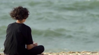 adam, kıvırcık saçlı Genç bir fırtınada denizin kenarında oturur ve dalgalar görünüyor.