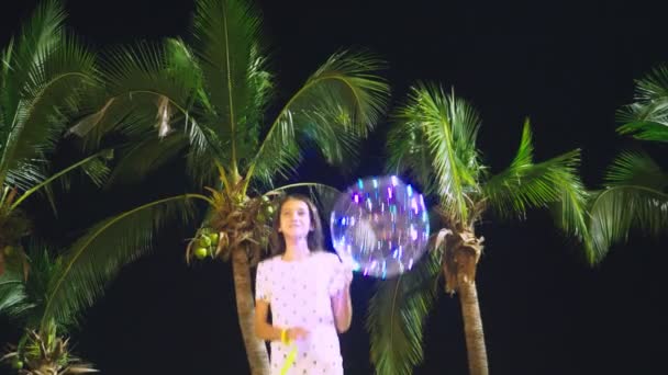 Happy teen girl holding big airy, glowing balloon in her hands against the background of palm trees. evening time. concept of tourism and beach holiday. — Stock Video