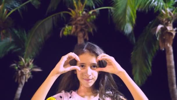 Kvinna visar hjärtat formen med fingrarna på bakgrunden av palmer på kvällen. — Stockvideo