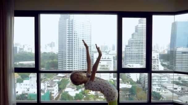 Delgada joven estirándose frente a una gran ventana panorámica con vistas a los rascacielos — Vídeos de Stock