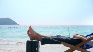 Genç başarılı iş adamı bir tembel turkuaz denizin karşı beyaz kumlu bir plaj üzerinde oturan ve diz üstü kullanarak. Bacaklarını bavula el bagajı ile yalan.