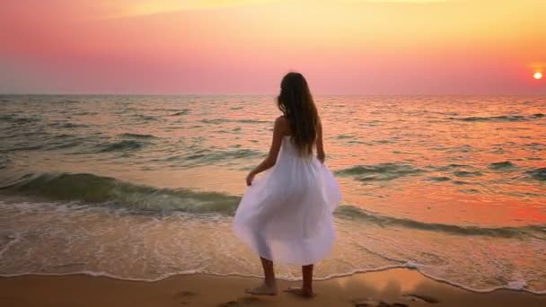 Chica adolescente en un largo vestido blanco con el pelo largo camina a lo largo de la orilla del mar al atardecer — Vídeos de Stock