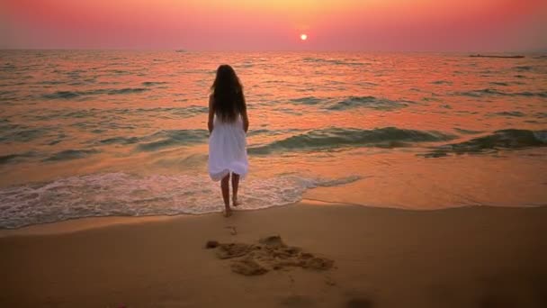 해질녘 해변을 따라 산책 하는 긴 머리를 가진 길고 흰 드레스 십 대 소녀 — 비디오