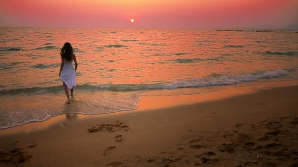Έφηβο κορίτσι σε ένα μακρύ άσπρο φόρεμα με μακριά μαλλιά περιπάτους κατά μήκος της ακτής στο ηλιοβασίλεμα — Αρχείο Βίντεο