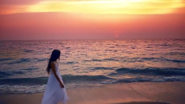 Het tiener meisje in een lange witte jurk met lang haar wandelingen langs de kust bij zonsondergang — Stockvideo