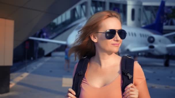 Het concept van het toerisme. Vrouw toerist in zonnebril met een rugzak op haar rug gaat naar de luchthaven op de achtergrond van het vliegtuig. vrouw blond glimlachen op de luchthaven — Stockvideo