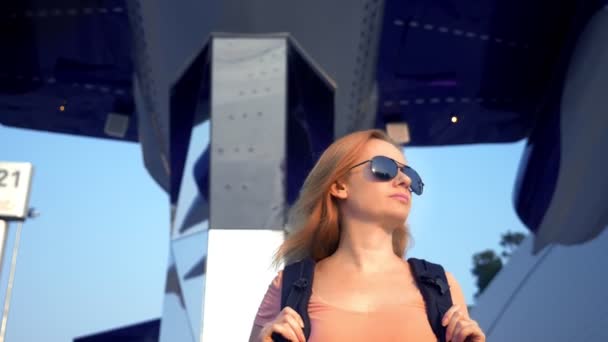 El concepto de turismo. Mujer turista en gafas de sol con una mochila en la espalda va al aeropuerto en el fondo del avión. mujer rubia sonriendo en el aeropuerto — Vídeos de Stock