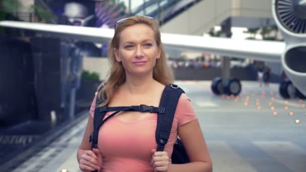Das Konzept des Tourismus. Eine Touristin mit Sonnenbrille und einem Rucksack auf dem Rücken geht im Hintergrund des Flugzeugs zum Flughafen. Blondine lächelt am Flughafen — Stockvideo
