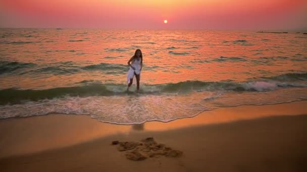 Adolescente menina em um vestido branco longo com longos passeios de cabelo ao longo da praia ao pôr do sol — Vídeo de Stock