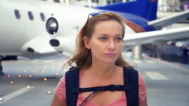 Das Konzept des Tourismus. Eine Touristin mit Sonnenbrille und einem Rucksack auf dem Rücken geht im Hintergrund des Flugzeugs zum Flughafen. Blondine lächelt am Flughafen — Stockvideo