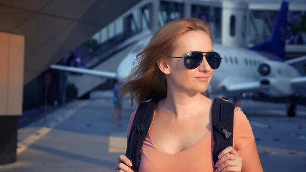 Turizmus fogalmának. Nő turista napszemüveg egy hátizsákot a hátán a repülőtér folyik a háttérben a gép. nő szőke, mosolygós a repülőtéren — Stock Fotó