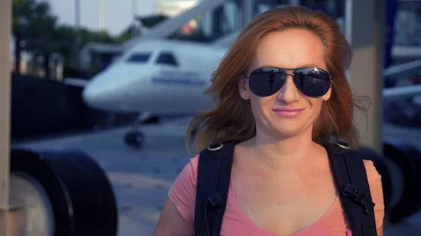 Het concept van het toerisme. Vrouw toerist in zonnebril met een rugzak op haar rug gaat naar de luchthaven op de achtergrond van het vliegtuig. vrouw blond glimlachen op de luchthaven — Stockfoto