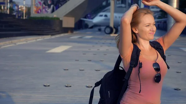 Het concept van het toerisme. Vrouw toerist in zonnebril met een rugzak op haar rug gaat naar de luchthaven op de achtergrond van het vliegtuig. vrouw blond glimlachen op de luchthaven — Stockfoto