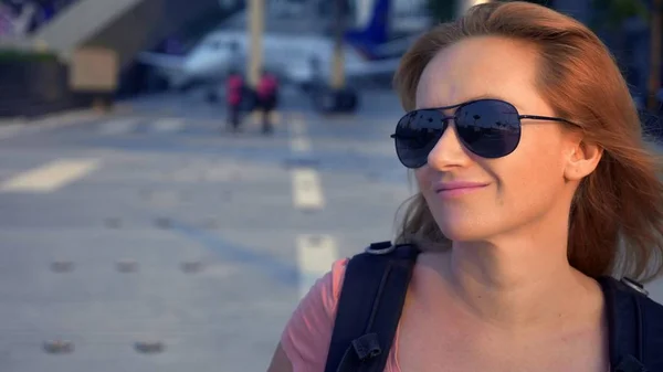 Koncepce cestovního ruchu. Žena turista v sluneční brýle s batohem na zádech přejde na letiště na pozadí letadla. žena blond se usmívá na letišti — Stock fotografie