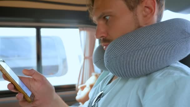 Un jeune homme avec un oreiller de voyage dans une voiture utilise un smartphone. Concept de voyage routier — Video