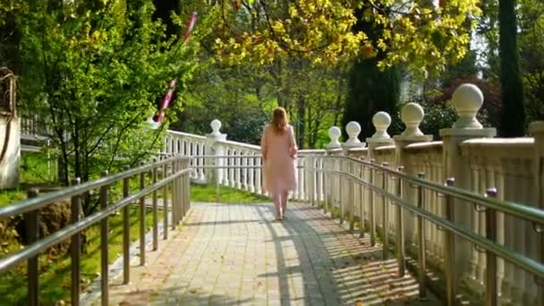 Una mujer con un abrigo rosa camina por un sendero con una balaustrada blanca en un parque tropical — Vídeo de stock