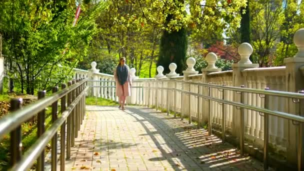 Una donna in cappotto rosa cammina lungo un sentiero con una balaustra bianca in un parco tropicale — Video Stock