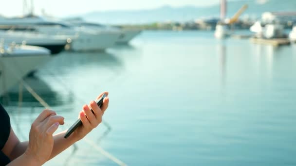 Mano femminile tiene smartphone su sfondo sfocato di porto con yacht — Video Stock
