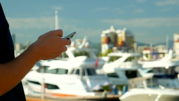 Mãos masculinas segurando smartphone no fundo borrado do porto com iates — Vídeo de Stock