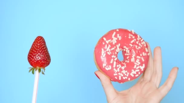 Концепция выбора между пончиком и клубникой. женские руки с красным лаком для ногтей держать с розовым пончиком и клубникой на палочке на синем фоне — стоковое видео