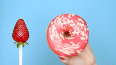 çörek ve çilek arasında seçim kavramı. mavi bir arka plan üzerinde bir sopa üzerinde pembe çörek ve çilek ile kırmızı oje tutun kadın eller