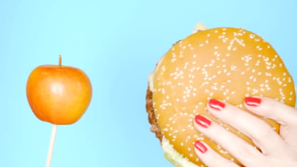 Concepto de comida saludable y poco saludable. Yaloko contra las hamburguesas sobre un fondo azul brillante. manos femeninas con esmalte de uñas rojo sosteniendo hamburguesa y manzana — Vídeos de Stock