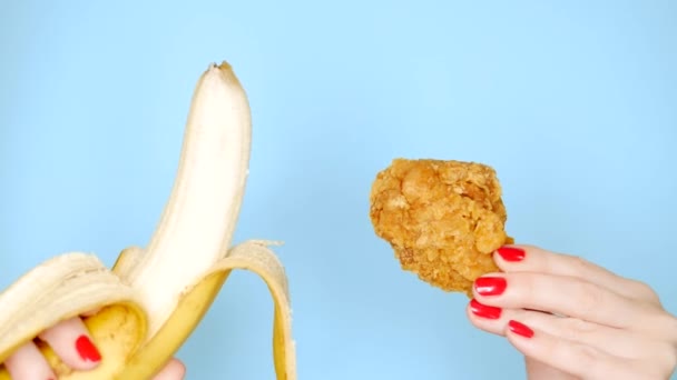 Conceito de alimentos saudáveis e insalubres. banana contra perna de frango assada frita em um fundo azul brilhante. mãos femininas com esmalte vermelho segurar banana e pepitas — Vídeo de Stock