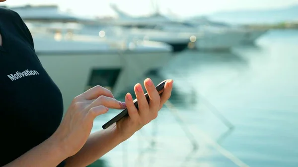 female hand holds smartphone on blurred background of port with yachts