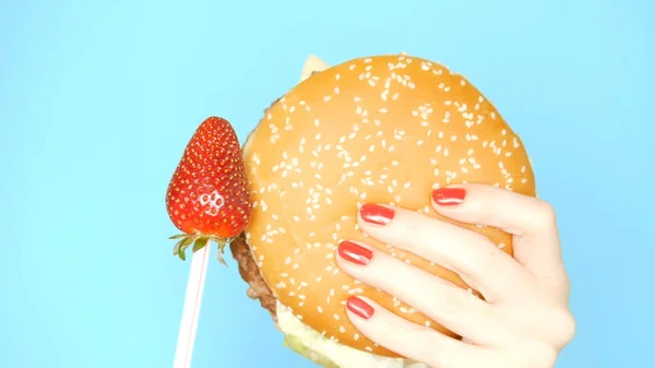 Conceito de alimentos saudáveis e insalubres. Morangos contra hambúrgueres em um fundo azul brilhante. mãos femininas com esmalte vermelho segurar hambúrguer e baga — Fotografia de Stock