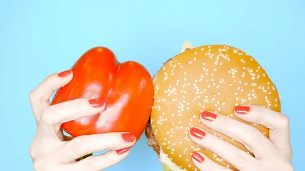 Sağlıklı ve sağlıksız gıda kavramı. parlak mavi bir arka plan üzerinde hamburger karşı tatlı kırmızı biber. kırmızı oje ile kadın elleri burger ve kırmızı biber tutun — Stok fotoğraf