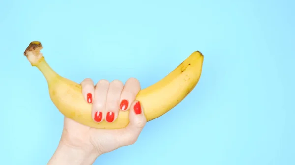 Frauenhände mit roten Nägeln halten eine gelbe Banane auf blauem Hintergrund — Stockfoto