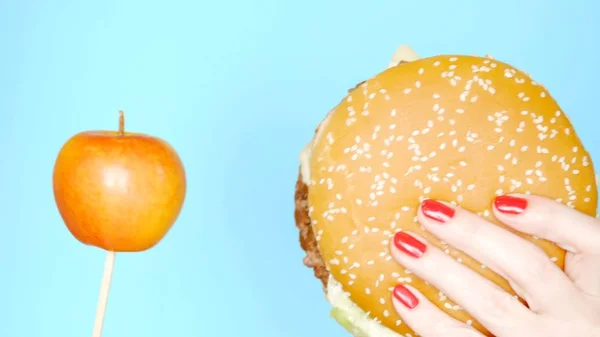 Conceito de alimentos saudáveis e insalubres. Yaloko contra hambúrgueres em um fundo azul brilhante. mãos femininas com esmalte vermelho segurando hambúrguer e maçã — Fotografia de Stock