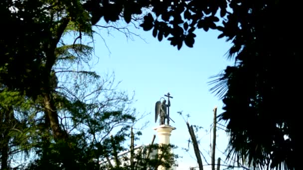 Suddig silhuett staty av en ängel med ett svärd mot den blå himlen i ramen för lövverk — Stockvideo