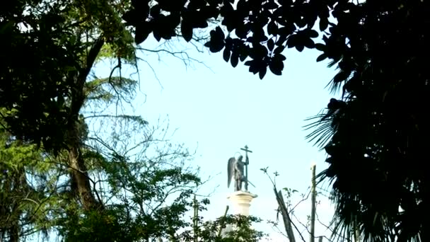 Verschwommene Silhouette Statue eines Engels mit einem Schwert gegen den blauen Himmel im Rahmen von Laub — Stockvideo