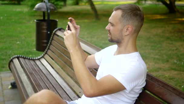 Piano medio. Un uomo blogger utilizza uno smartphone mentre è seduto su una panchina del parco. Ha una videochiamata con qualcuno. — Video Stock