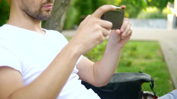 クローズ アップ。男性ブロガーは、公園のベンチに座っている間、スマートフォンを使用しています。彼は誰かとビデオ通話を持っている — ストック動画