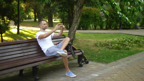 Πλήρης βολή. Ένας άνθρωπος blogger χρησιμοποιεί ένα smartphone, ενώ κάθονται σε ένα παγκάκι πάρκο. Έχει βιντεοκλήση με κάποιον — Αρχείο Βίντεο
