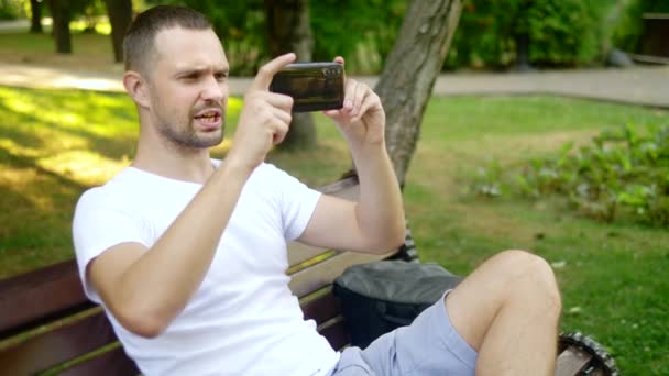 Plan medio. Un bloguero hombre utiliza un teléfono inteligente mientras está sentado en un banco del parque. Tiene una videollamada con alguien — Vídeos de Stock